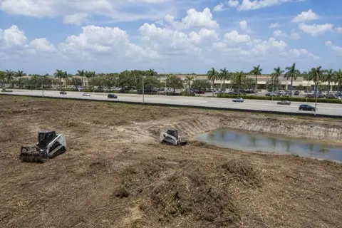 Broward County commercial parcel mid-clearing with windrowed mulch, perimeter silt fencing, and a started retention-pond dig